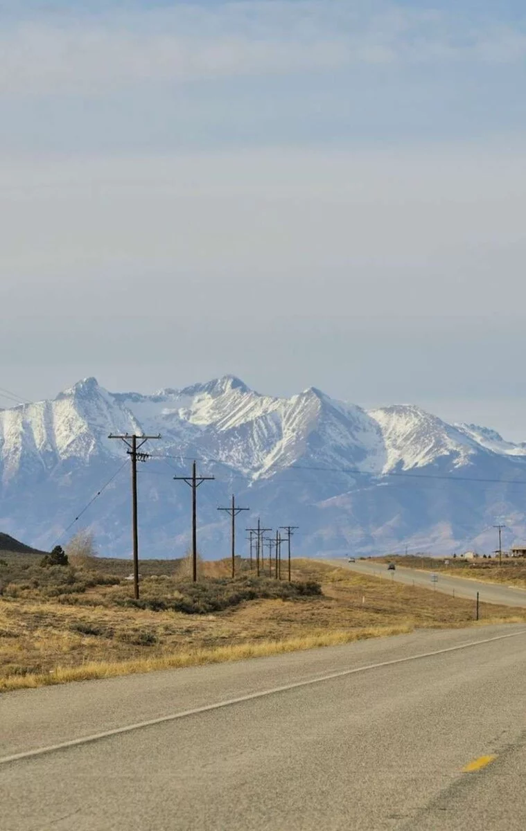 A quiet rural highway in Montana lined with utility poles and backed by snow-covered peaks—representing fuel logistics in remote farm regions.