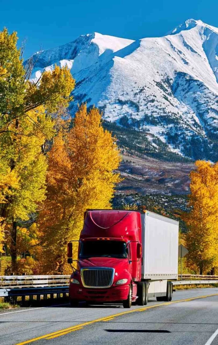 A red semi-truck drives through Montana’s autumn landscape with golden trees and snow-capped mountains, symbolizing rural fuel transport.