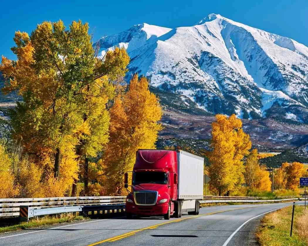 A red semi-truck drives through Montana’s autumn landscape with golden trees and snow-capped mountains, symbolizing rural fuel transport.