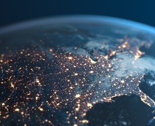 Aerial view of Earth at night, showing glowing city lights across continents and a curved horizon against a dark sky.