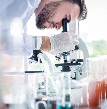 Scientist closely examining a sample through a microscope while wearing gloves in a bright laboratory setting.