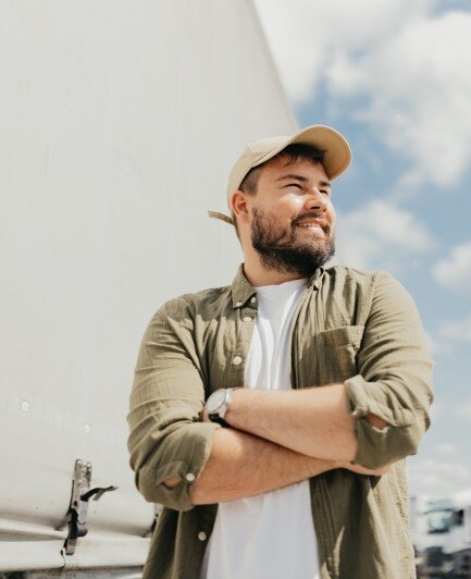 Man smiling with arms crossed while standing outside on a sunny day, wearing a cap and casual shirt with a bright sky behind him.
