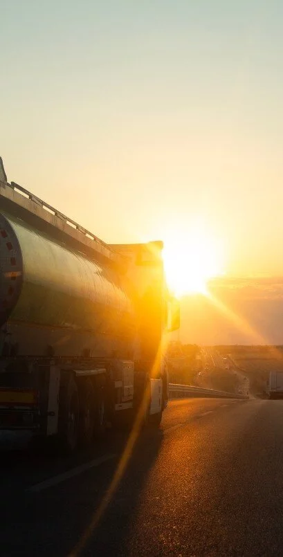 Tanker truck driving along a highway at sunset, with warm sunlight reflecting off the trailer and the road ahead.